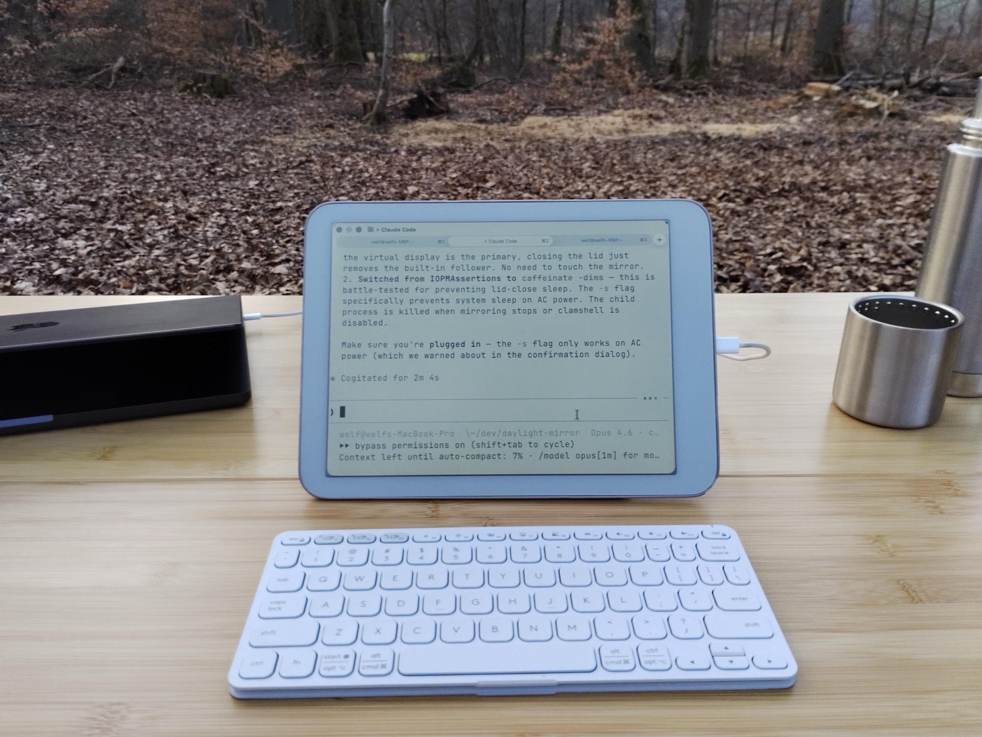 Daylight DC-1 on MacBook showing Claude Code, apples and water bottle on bamboo table in forest
